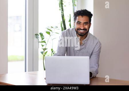 Ein lächelnder Mann mit Bart an einem Holztisch in einem gut beleuchteten Raum, der an einem Laptop arbeitet Stockfoto