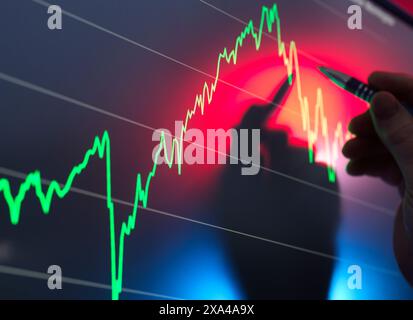 Eine Person untersucht Finanzdaten auf einem Bildschirm, auf dem ein Diagramm mit aufwärts- und abwärts gerichteten Trends angezeigt wird, und verwendet einen Stift, um das Liniendiagramm zu verfolgen. Stockfoto