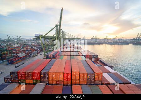 Ein Blick aus der Vogelperspektive auf ein Frachtschiff mit bunten Containern, das während Sonnenuntergang an einem geschäftigen Hafen angedockt ist. Stockfoto