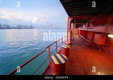 Das Bild zeigt vom Deck eines Schiffes aus einen lebendigen Blick mit roten und gelben Geländern, ruhigem Meerwasser und Containerkränen an einem Hafen in der Ferne unter teilweise bewölktem Himmel. Stockfoto