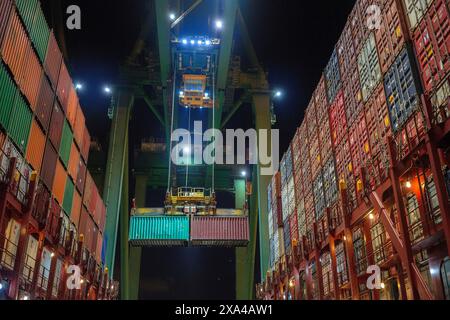 Ein Containerschiff wird nachts in einem Hafen be- oder entladen, wobei ein Portalkran einen Container in mehrfarbigen Containern bewegt. Stockfoto