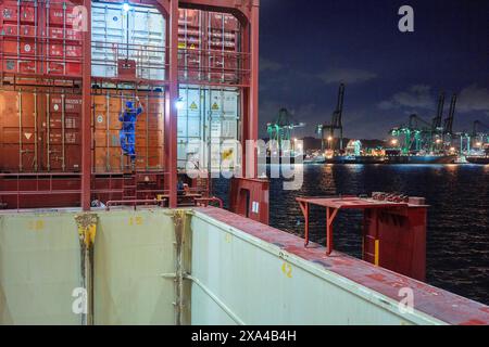 Ein Frachtschiff legt nachts an einem belebten Hafen an, wobei Container auf Deck gestapelt sind und Kräne im Hintergrund sichtbar sind. Stockfoto