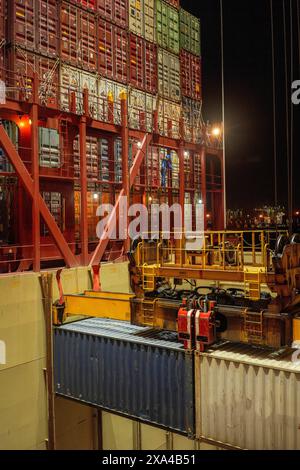 Nächtlicher Blick auf einen belebten Containerhafen, bei dem ein Schiff von einem großen, beleuchteten Portalkran mit farbenfrohen Containern be- oder entladen wird. Stockfoto