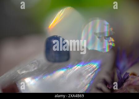 Ein Nahaufnahme, verschwommenes Bild eines Kristalls mit einem Prismeneffekt, der Regenbogenlichtbrechungen erzeugt, begleitet von einem undeutlichen blauen Objekt und Pflanzenmaterial. Stockfoto