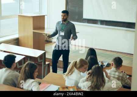 Interaktive Lernsitzung. Junger Lehrer leitet Vorlesung vor dem Klassenzimmer, wobei er sich mit verschiedenen Schülergruppen auseinandersetzt. Stockfoto