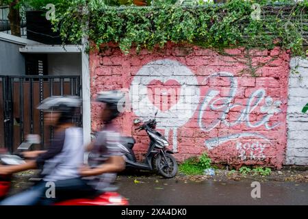 Motorradfahrer fahren an einer leuchtend roten Wand vorbei, auf der ein Herz und das Wort „Liebe“ unter einem üppig grünen Blätterdach gemalt sind. Stockfoto