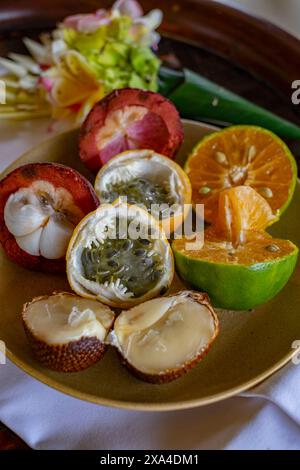 Ein Teller mit einer Auswahl an tropischen Früchten, darunter geschnittene Orangen, Passionsfrüchte und andere, serviert auf einem Holztablett mit bunten Blumen im Hintergrund. Stockfoto