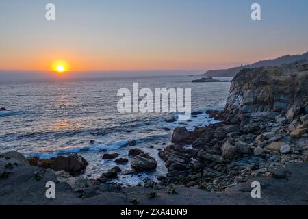 Das Bild fängt einen ruhigen Sonnenuntergang über einer zerklüfteten Küste mit Wellen ein, die sanft auf die Felsen darunter stürzen. Stockfoto