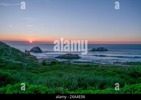 Blick auf den Sonnenuntergang über eine Küstenlandschaft mit hügeligen Hügeln, üppigem Grün im Vordergrund und mehreren Felsformationen im Ozean. Stockfoto