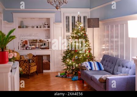 Ein gemütliches Wohnzimmer, das zu Weihnachten dekoriert ist, mit einem beleuchteten Weihnachtsbaum, einem grauen Sofa und einer Küche im Hintergrund. Stockfoto
