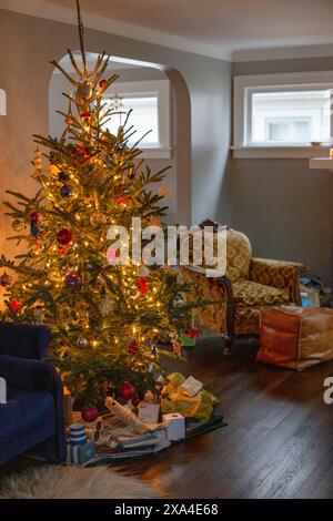 Ein warm beleuchtetes Wohnzimmer ist mit einem dekorierten Weihnachtsbaum mit bunten Lichtern und verschiedenen Ornamenten geschmückt, umgeben von geöffneten Geschenkboxen und gemütlichen Stühlen in der Nähe. Stockfoto