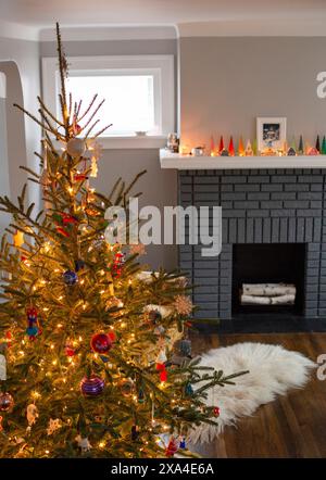 Ein gut dekorierter Weihnachtsbaum steht in einem gemütlichen Raum mit einem beleuchteten Kamin und einer Reihe von beleuchteten Kerzen auf dem Kaminsims. Stockfoto