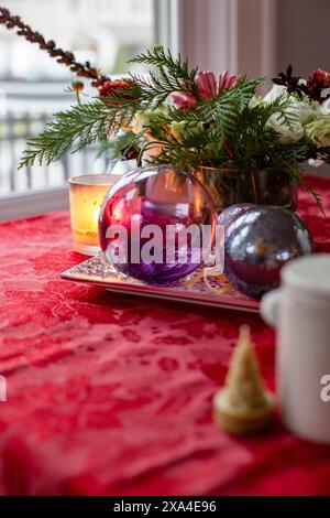 Ein festlicher Tisch mit einer leuchtend roten Tischdecke, die ein dekoratives Arrangement aus Blumen, Kerzen und bunten Kugeln auf einem silbernen Tablett neben einer weißen Kanne und einem kleinen dekorativen Baum zeigt. Stockfoto