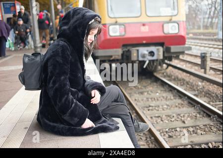 Eine Person in einem schwarzen Kapuzenmantel sitzt auf einem Bahnsteig, blickt auf ihre Uhr, mit einem Zug und Menschen im Hintergrund. Stockfoto