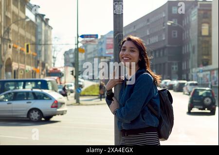 Eine lächelnde Frau mit einem Rucksack, die auf einer Straße steht und sich an einem sonnigen Tag an einem Wegweiser lehnt. Stockfoto