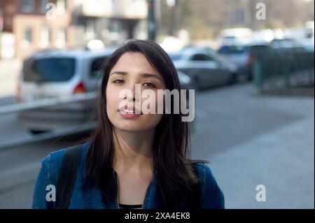 Eine Frau mit langen braunen Haaren und einer Jeansjacke steht draußen mit Fahrzeugen und Gebäuden im Hintergrund und blickt mit einem leichten Lächeln in die Kamera. Stockfoto