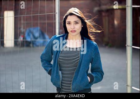 Eine junge Frau steht vor einem Maschendrahtzaun, mit dunklen Haaren, die im Wind wehen; sie trägt eine blaue Jacke über einem gestreiften Oberteil. Stockfoto