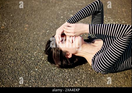 Eine Frau liegt auf dem Rücken auf dem Boden, lacht mit ihren Händen, die teilweise ihr Gesicht bedecken, und trägt ein gestreiftes Top. Stockfoto