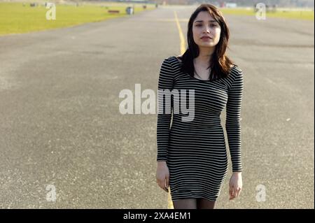 Eine Frau steht auf einer leeren Straße mit Grasfeldern im Hintergrund und trägt ein schwarz-weiß gestreiftes Kleid. Stockfoto