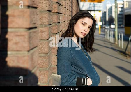 Eine Frau lehnt sich an eine Ziegelmauer mit urbanem Hintergrund und blickt tagsüber entspannt auf die Kamera. Stockfoto