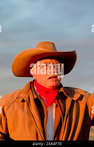 Ein älterer Mann mit Brille und Schnurrbart mit Cowboyhut und rotem Halstuch schaut in die Ferne. Er trägt eine braune Jacke, und der Hintergrund deutet auf Sonnenuntergang hin. Stockfoto