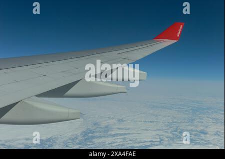 Ein Flugzeugflügel mit roter Spitze schwingt über den Wolken und der eisbedeckten Landschaft darunter und fängt die Majestät des Fluges in einem klaren blauen Himmel ein. Stockfoto