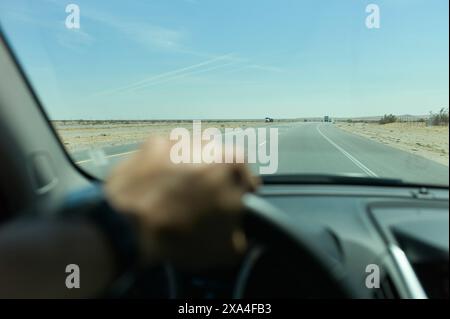 Die Hand des Fahrers am Lenkrad aus Sicht des Innenraums eines Autos, mit freier Sicht auf eine gerade Straße und einen blauen Himmel. Stockfoto