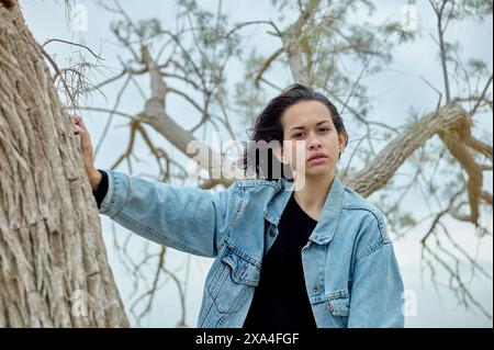 Eine Person mit schulterlangen Haaren steht draußen, lehnt sich lässig an einen Baum, trägt eine Jeansjacke über einem schwarzen Top, mit einem ernsten Gesichtsausdruck und einem kargen Baum und bewölktem Himmel im Hintergrund. Stockfoto