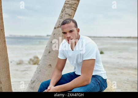 Ein Mann sitzt an einem Baumstamm auf sandigem Boden und blickt mit heiterem Ausdruck in die Kamera, mit klarem Himmel und flachem Horizont im Hintergrund. Stockfoto