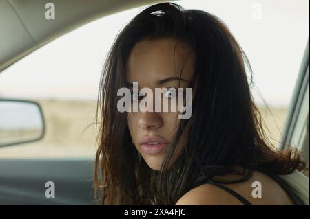 Eine Frau sitzt in einem Auto, dreht sich um, um die Kamera mit einem intensiven Blick zu betrachten, ihr dunkles Haar zerzaust und einen absichtlichen, nachdenklichen Gesichtsausdruck. Stockfoto