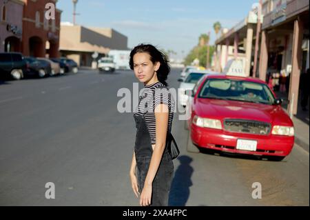Eine Frau steht selbstbewusst in einer Stadtstraße mit Geschäften im Hintergrund und einem roten Auto, das an der Seite geparkt ist. Stockfoto