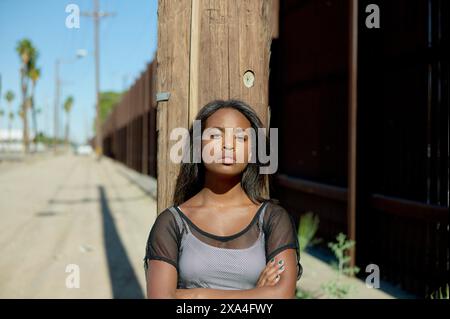 Eine junge Frau steht selbstbewusst in einer Straßenlage, lehnt sich mit überkreuzten Armen an einen Holzmast. Stockfoto