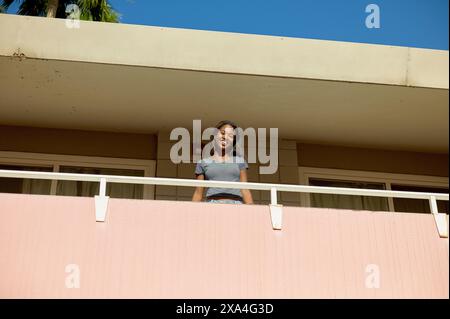Eine fröhliche junge Frau steht auf einem Balkon mit einer rosa Wand, auf einem weißen Geländer vor einem klaren blauen Himmel. Stockfoto