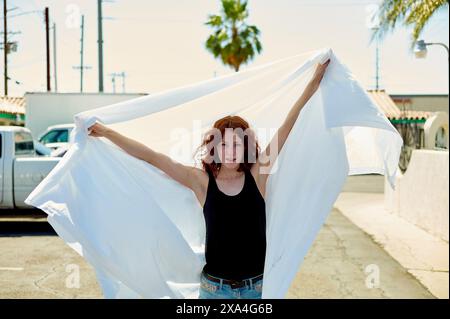 Eine Frau steht auf einer sonnigen Straße und hält einen großen, durchsichtigen weißen Stoff über dem Kopf, der in der Brise weht. Sie trägt ein schwarzes ärmelloses Oberteil und blaue Jeans. Stockfoto