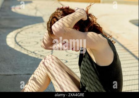 Eine Frau, die draußen auf dem Boden sitzt, schirmt ihr Gesicht mit ihrem Arm vor der Sonne ab. Stockfoto