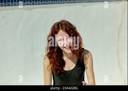 Eine fröhliche junge Frau mit langen roten Haaren steht neben einem Pool. Ihr strahlendes Lächeln deutet auf einen Moment der Freude oder des Lachens im hellen Sonnenlicht hin. Stockfoto