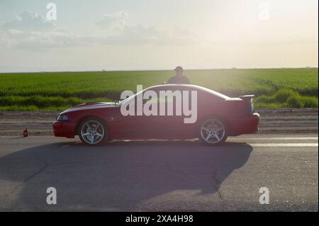 Eine Person steht durch das Sonnendach eines roten Sportwagens am Straßenrand mit üppigen grünen Feldern und einem klaren Himmel im Hintergrund. Stockfoto