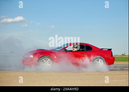 Ein roter Sportwagen führt einen Burnout durch, wobei weißer Reifenrauch aus den Hinterreifen bläst und ein Fahrer durch die Seitenscheibe sichtbar ist. Stockfoto