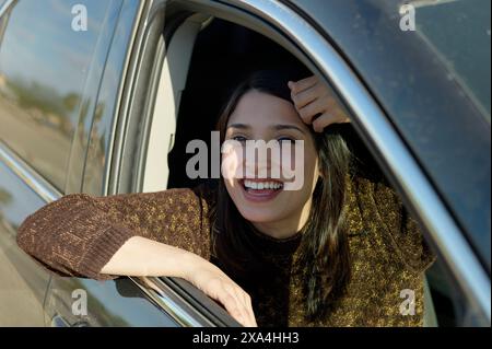 Eine lächelnde Frau lehnt sich aus dem Fenster eines Autos auf der Fahrerseite, ihre Hand liegt auf dem Kopf, während sie einen sonnigen Tag genießt. Stockfoto