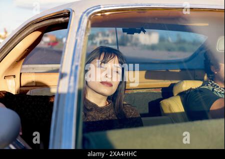Eine Frau mit dunklen Haaren sitzt auf dem Beifahrersitz eines Oldtimers und blickt mit einem nachdenklichen Ausdruck aus dem Fenster, während der Fahrer teilweise sichtbar ist und sich auf die Frau konzentriert. Stockfoto