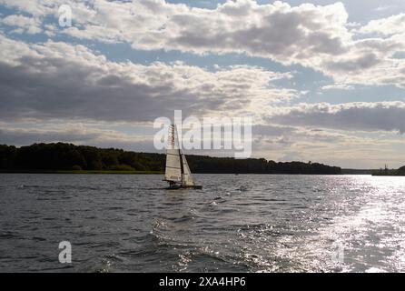 Ein Segelboot gleitet über das Wasser, mit einer bewaldeten Küste in der Ferne, unter einem Himmel voller weißer Wolken. Stockfoto