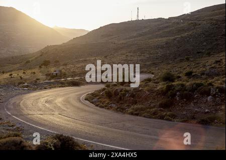 Eine gewundene Straße schlängelt sich bei Sonnenuntergang durch eine hügelige Landschaft, mit warmem Licht, das lange Schatten und ein goldenes Leuchten über die Szene wirft. Stockfoto