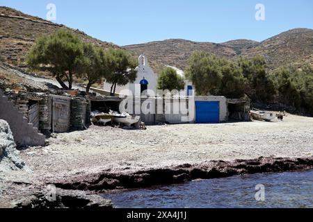 Eine ruhige Küstenszene mit einer kleinen weißen Kirche mit einer blauen Kuppel vor einer hügeligen Landschaft mit umliegenden Sträuchern. Boote und Bauwerke befinden sich in der Nähe des Wasserrandes unter einem hellblauen Himmel. Stockfoto