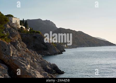 Eine ruhige Küstenlandschaft mit einer zerklüfteten Küste mit Gebäuden auf den Klippen, mit Blick auf das ruhige Meer mit Bergen im Hintergrund in der Dämmerung. Stockfoto