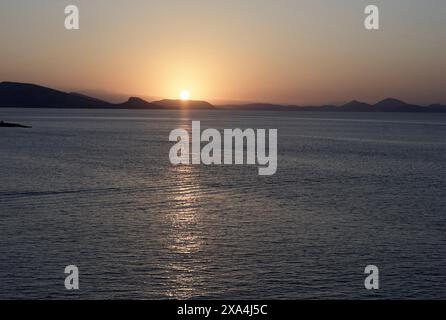 Ein ruhiger Sonnenuntergang über dem Meer mit der Sonne, die ein warmes Leuchten ausstrahlt, und ein reflektierender Pfad auf dem Wasser, eingerahmt von fernen Silhouetten von Bergen. Stockfoto