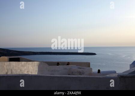Silhouetten von Individuen sind über einer weiß getünchten Wand zu sehen, mit Blick auf ein ruhiges Meer in der Dämmerung, mit klarem Himmel und einem ruhigen Horizont. Stockfoto