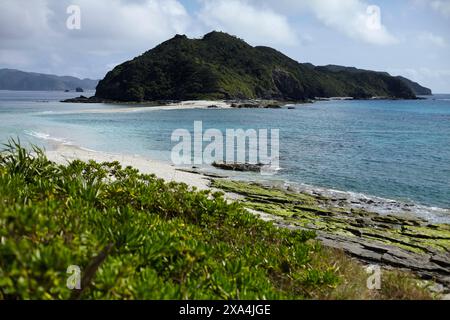 Ein ruhiger Strand mit weißem Sand liegt eingebettet zwischen üppigen grünen Hügeln unter einem klaren blauen Himmel, mit Felsformationen, die in das türkisfarbene Meer ragen. Stockfoto