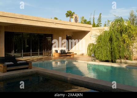 Ein modernes Haus mit großen Glasfenstern überblickt einen ruhigen Swimmingpool, umgeben von üppiger Vegetation und einem klaren blauen Himmel. Stockfoto