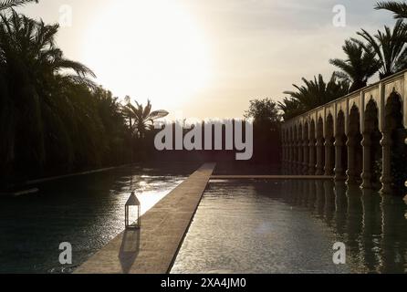 Das Bild zeigt einen ruhigen Swimmingpool in der Abenddämmerung, in dem das Sonnenlicht durch Palmen strömt und von der Wasseroberfläche reflektiert wird, neben einer kunstvollen Balustrade. Stockfoto