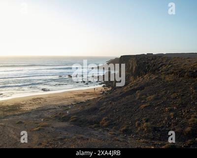 Der Sonnenaufgang beleuchtet eine ruhige Strandlandschaft mit einer zerklüfteten Klippe entlang des Ozeans, die sanftes Licht auf Sand und Felsen wirft. Stockfoto